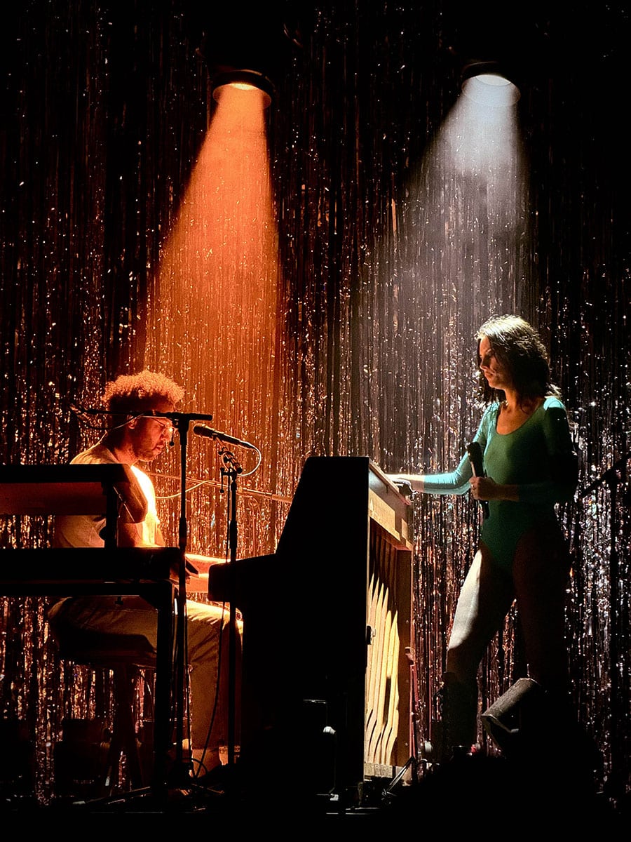 This picture shows a concert photo of the band Wolf Alice, taken by Sara Vukman. The photo features lead singer performing on stage with white light on her and the orange light is on the piano player.